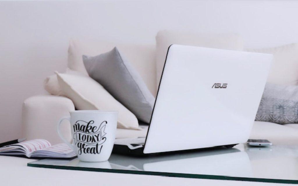 Photographie d'un ordinateur sur une table basse avec une tasse de café et en fond un canapé orné de coussins